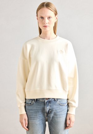 Felpa color crema con scollo a costine e polsini. Presenta un fit cropped e un logo ricamato sottile sul petto. Abbinata a jeans azzurri chiari.