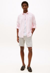 Hellrosa Hemd über einem weißen T-Shirt, kombiniert mit grauen Shorts und schwarzen Slippers. Glatte Stoffqualität und lässige Passform.