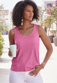 Roze getextureerde tanktop met wijde halslijn, gecombineerd met witte broek. Persoon houdt een lichtblauw koffie kopje vast, draagt kralenarmbanden en franjesoorbellen.