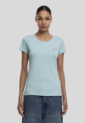 Femme portant un t-shirt bleu clair avec un petit contour de visage de chien noir sur le côté gauche de la poitrine, associé à un jean bleu.