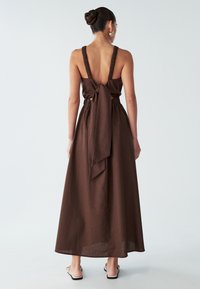 Sieviete ar matiem bunā, ģērbtā brūnā bez piedurknēm maksi kleitā ar savilktu jostu un lielu loku aizmugurē, stāvot uz gaiša fona.