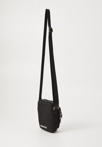 Černá crossbody taška vyrobená z hladké látky, s uzavíráním na zip, nastavitelným popruhem a stříbrnou logem vzadu.