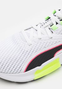 Zapatilla deportiva blanca con parte superior de malla texturizada, acentos en negro y rosa, detalles en verde neón y una suela acolchada visible.