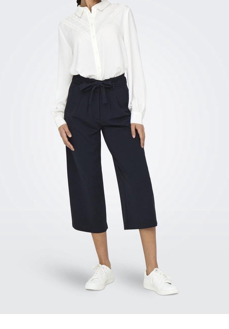 Femme portant un chemisier blanc à manches longues rentré dans un pantalon noir à jambes larges et taille haute, coupé court, avec une ceinture nouée et des baskets blanches.