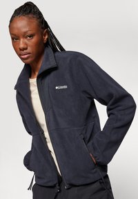 Dunkelblaues Fleece-Jacke mit hohem Kragen, Reißverschlusstaschen und dezentem Logo auf der Brust. Weicher Stoff, durchgehender Reißverschluss für einfache Handhabung.