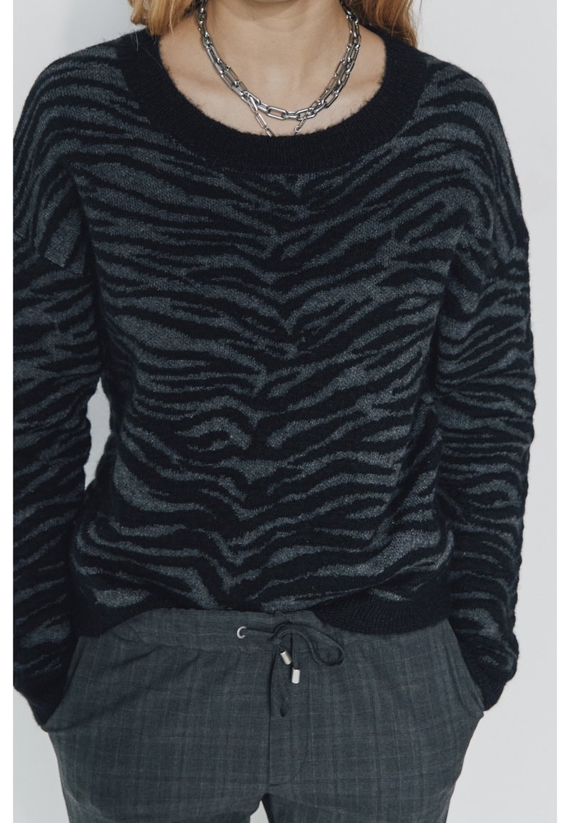 Un pull noir présentant un motif zèbre en noir et gris, avec un décolleté large et des poignets côtelés. Associé à un pantalon texturé gris.