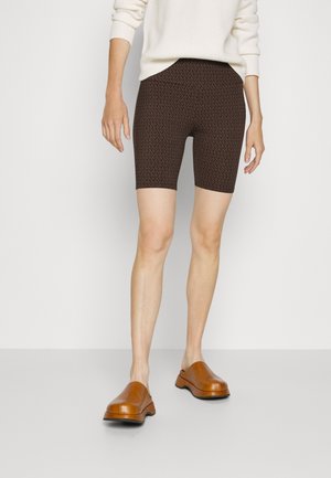 Person iført brune mønstrede biker shorts, hvid langærmet sweater og brune slip-on sko, stående foran en ensfarvet baggrund.