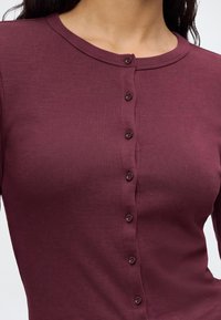 Gros plan d'une personne portant une chemise côtelée bordeaux à manches longues, boutonnée avec de petits boutons ronds sur le devant.