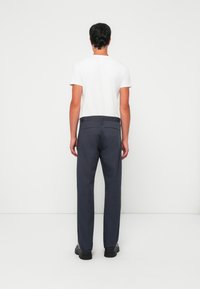 Uomo in piedi di spalle, che indossa una maglietta bianca a maniche corte, pantaloni blu scuro e scarpe nere su uno sfondo bianco uniforme.