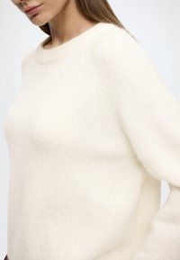 Krém színű kötött pulóver puha, texturált felülettel, bordázott kerek nyakkivágással és széles raglán ujjal.