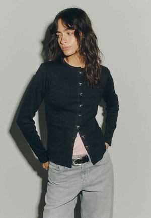 Young woman with wavy dark hair wears a black long-sleeve top with small bow fasteners over a pink shirt and light gray jeans.