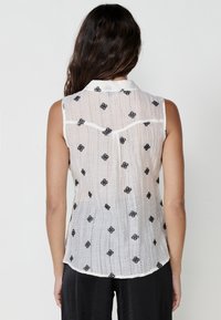 Bluz�ă albă fără mâneci cu modele florale negre brodate, țesătură texturată și un design subtil cu dungi. În spate, cu guler.