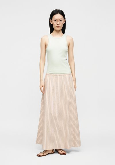 Femme aux longs cheveux noirs portant des lunettes transparentes, un haut vert clair sans manches, une jupe longue rayée beige et des sandales marron, debout.