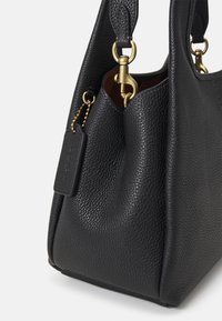 Czarna teksturowana skórzana torebka z złotymi zapięciami i małą wiszącą metką na jasnym tle, pokazana z boku w zbliżeniu.