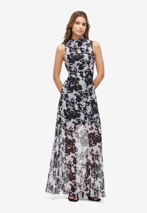 Kvinne står med hendene bak ryggen iført ermeløs, gulvlengde svart og grå mønstret kjole og svarte åpne hæler på hvit bakgrunn.