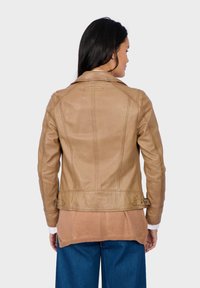 Braune Lederjacke mit tailliertem Schnitt, ausgestattet mit einem strukturierten Kragen, vertikalen Nähten und verstellbaren seitlichen Schnallen. Über einem hellen Pullover getragen.