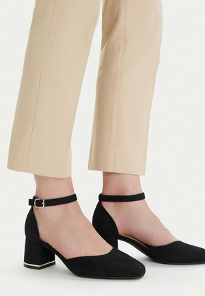 Tacones de bloque de ante negro con tiras en el tobillo, usados con pantalones cortos beige sobre fondo claro.