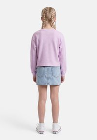 Dítě s blonďatými copánky, které má na sobě lila svetr, světle modrou džínovou sukni a bílé vysoké tenisky, stojící zády.