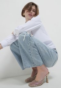 Jeans azzurri a gamba larga con orlo sfilacciato, abbinati a mule con tacco dorati lucidi caratterizzati da un motivo a rete metallica. Maglietta bianca a maniche lunghe.