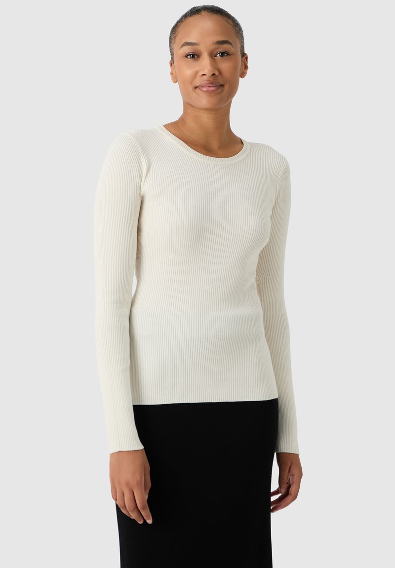 Weißes, geripptes Langarmshirt mit rundem Ausschnitt, taillierter Form und weicher Textur, kombiniert mit einem schwarzen Rock, Modell schaut nach vorne.