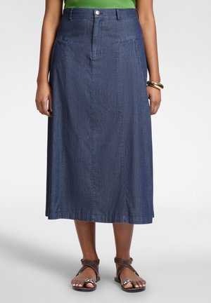 Femme portant une jupe en denim bleue longueur midi avec poches frontales et fermeture à boutons, associée à des sandales marron à lanières et un haut vert.
