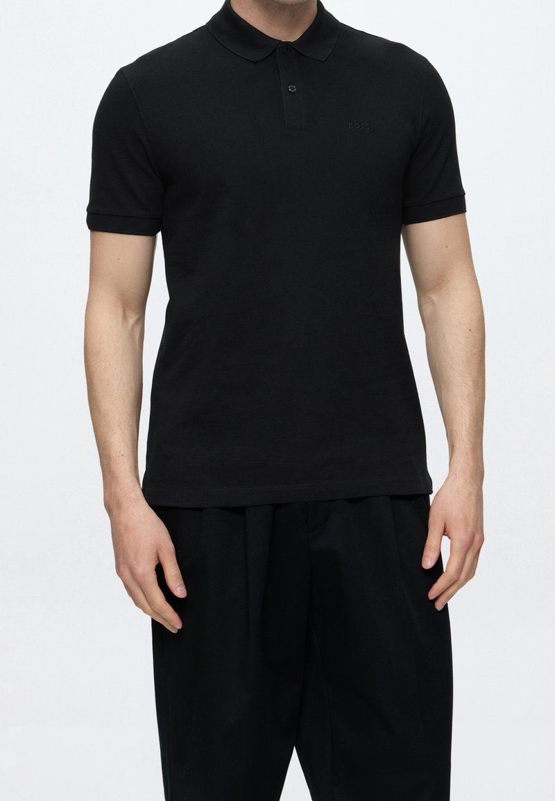 Homme portant un polo noir ajusté à manches courtes et un pantalon noir ample sur un fond clair uni.