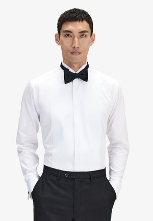 White tuxedo shirt with vertical pleats, black bow tie, button cuffs, and a structured collar. Worn with black trousers, showcasing a formal look.