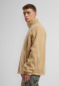 Pulover bej din fleece cu guler înalt, suprafață texturată și mâneci lungi. Designul prezintă o croială lejeră și detalii de cusătură vizibile.