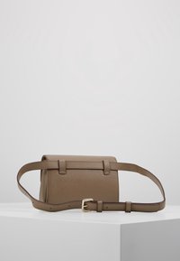 Sac à main en cuir marron avec un design structuré, doté d'une poignée courte, d'une bandoulière réglable et de coutures discrètes, posé sur une surface blanche.