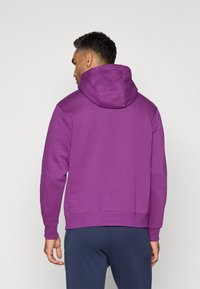 Lilla hættetrøje med ribbet talje og manchetter. Blødt stof, oversized hætte. Tætsiddende design, båret med marineblå bukser.