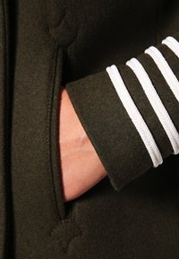 Abrigo de lana verde oscuro con un bolsillo lateral y un ribete trenzado blanco en los puños, que presenta una textura suave y un diseño ajustado.