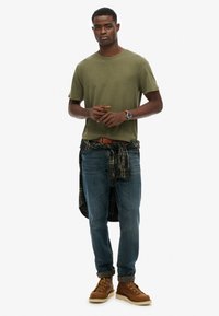 Zielona oliwkowa koszulka, niebieskie dżinsy, brązowe sznurowane buty oraz koszula w kratę związana w talii. Na sobie brązowy pasek i niebieski zegarek.