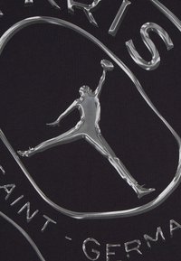 Μαύρο ύφασμα με λαμπερό, ασημί ανάγλυφο λογότυπο που παρουσιάζει μια φιγούρα που πηδάει και το κείμενο "PARIS SAINT-GERMAN", με ομαλές, αντανακλαστικές λεπτομέρειες.
