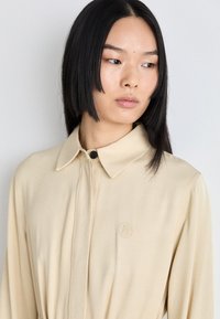 Béžová košeľa s golierom vyrobená z hladkej látky, s predným gombíkom a vyšitým logom, s dlhými rukávmi a voľným strihom.