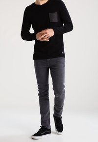 Svart långärmad skjorta med en grå ficka, ihop med mörkgrå slim-fit jeans och svarta sneakers, mot en vit bakgrund.