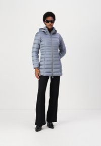 Giacca puffer grigia chiara con cappuccio, caratterizzata da cuciture orizzontali, una cerniera frontale e tasche laterali. Indossata con pantaloni neri e stivali.