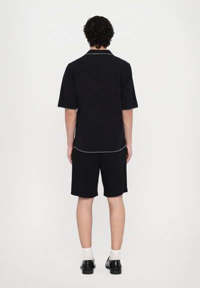 Black short-sleeve shirt with contrasting white stitching, paired with black shorts. The fabric appears textured; minimalistic design.