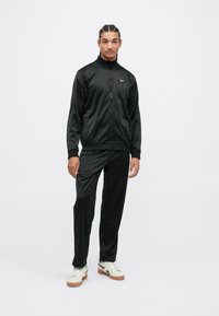 Vyras su pynėmis, apsirengęs juoda zip-up sportine striuke ir atitinkančiomis kelnėmis, stovi su baltais ir rudi batais ant balto fono.