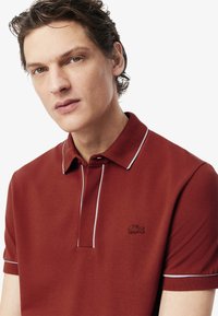 Röd polo skjorta i strukturerad tyg med krage, korta ärmar och tunna vita detaljer längs kragen och knappraden.