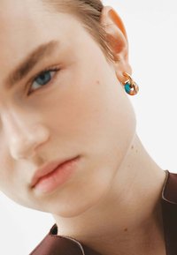 Pendiente de aro dorado con un acento de resina azul y naranja, diseño circular con textura suave, llevado en la oreja izquierda.