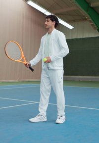 Fato de treino atlético branco com um casaco de fecho e calças combinando, segurando uma raquete de ténis laranja e uma bola de ténis verde num court azul.