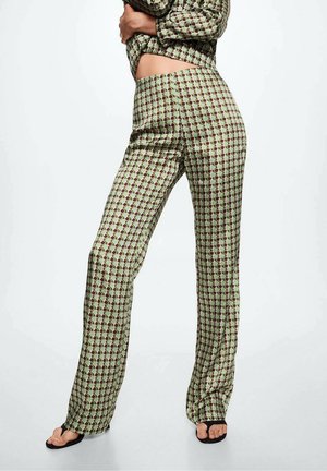Personne portant un pantalon taille haute à motifs et une chemise à manches longues assortie, debout avec un bras sur le ventre, sandales noires à bout ouvert visibles.