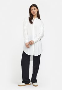 Camicia bianca con colletto, realizzata in tessuto lucido, con maniche lunghe, chiusura frontale a bottoni e orlo curvo, abbinata a pantaloni neri larghi e scarpe bianche.