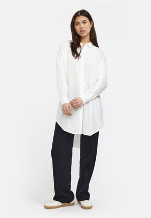 Camicia bianca con colletto, realizzata in tessuto lucido, con maniche lunghe, chiusura frontale a bottoni e orlo curvo, abbinata a pantaloni neri larghi e scarpe bianche.