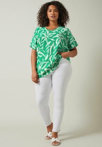 Top verde de manga corta con un patrón abstracto de hojas blancas, diseño de escote en V, combinado con jeans blancos ajustados y sandalias blancas tipo slide.