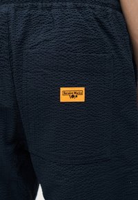 Παντελόνια από υφασμάτινο ύφασμα ναυτικού μπλε με τετράγωνη πίσω τσέπη που διαθέτει πορτοκαλί ετικέτα με την επιγραφή "Service Works" και μικρά εικονίδια εργαλείων.