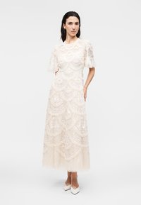 Γυναίκα που στέκεται φορώντας λευκό δαντελένιο φόρεμα μήκους μέσης με κυματιστά σχέδια και φουσκωτά μανίκια, συνδυασμένο με λευκά μυτερά τακούνια.