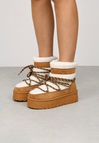 Botas marrón claro y crema con un upper texturizado que imita piel de animal, atadas con cordones de cuerda marrones, y con una suela gruesa y estriada.