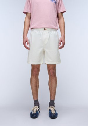 Mand iført lyserød t-shirt, hvide elastiske shorts med knap, grå sokker og blå sneakers står mod en ensfarvet baggrund.