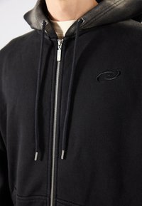 Felpa nera con zip, dal tessuto morbido, con cappuccio bicolore, cordini e logo ricamato sul petto.